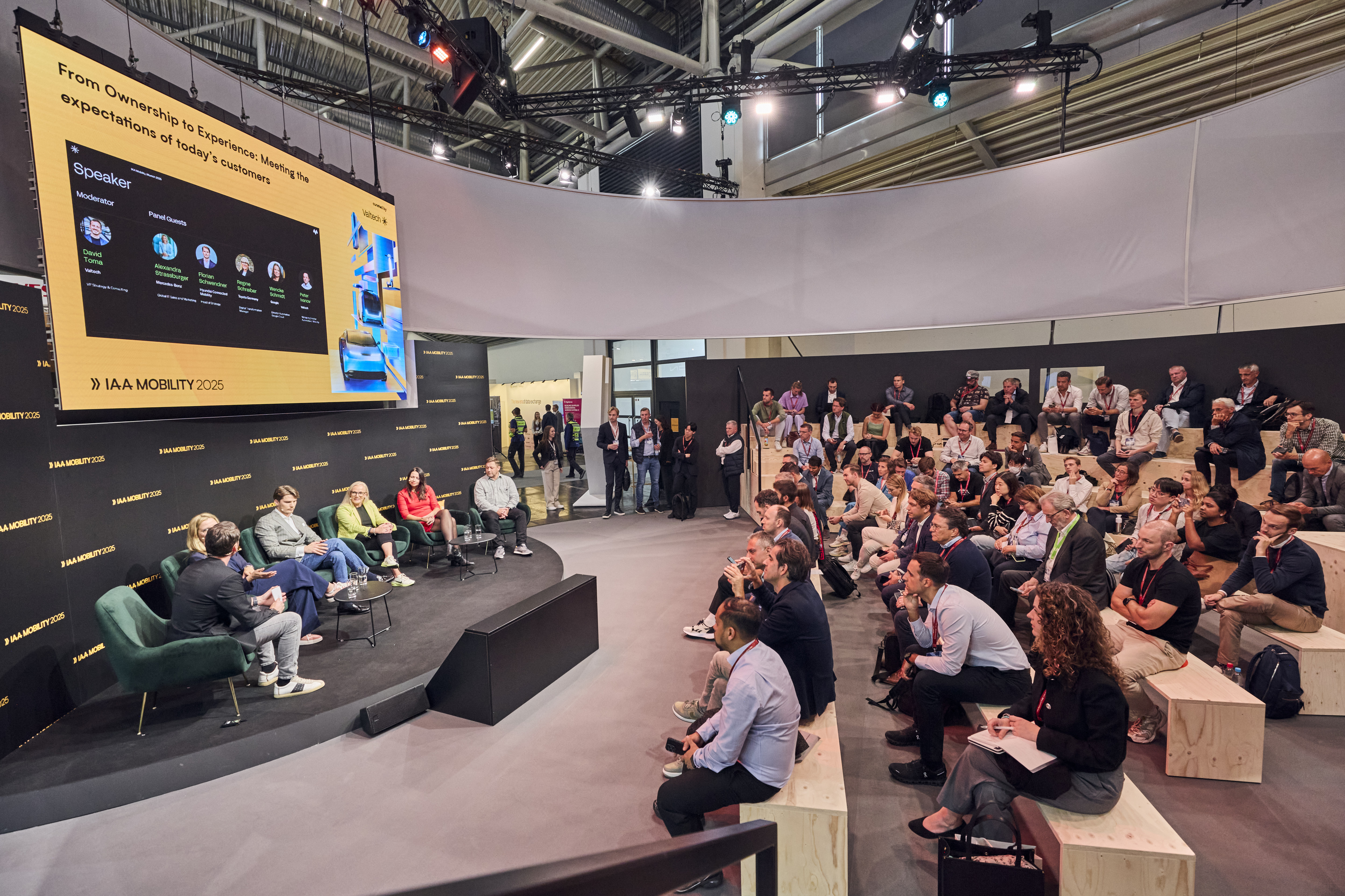 A large crowd sits on wooden benches, watching a presentation on a big screen in an outdoor setting.  