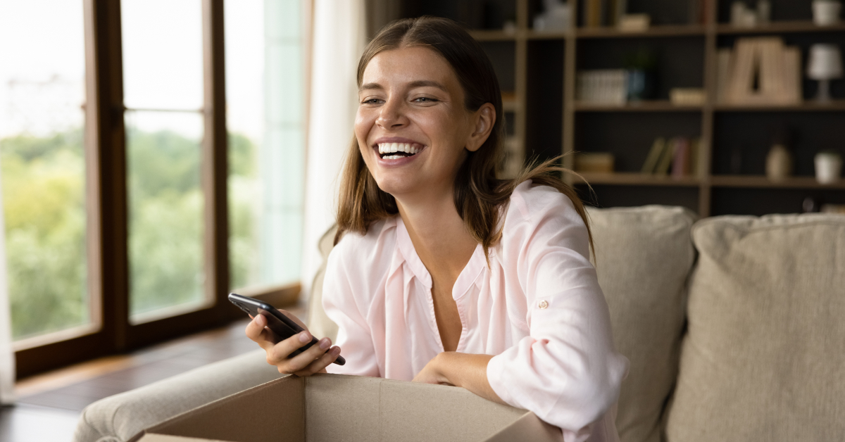 A woman smiles joyfully while holding her phone and unboxing a package, having ordered it online on a commerce website