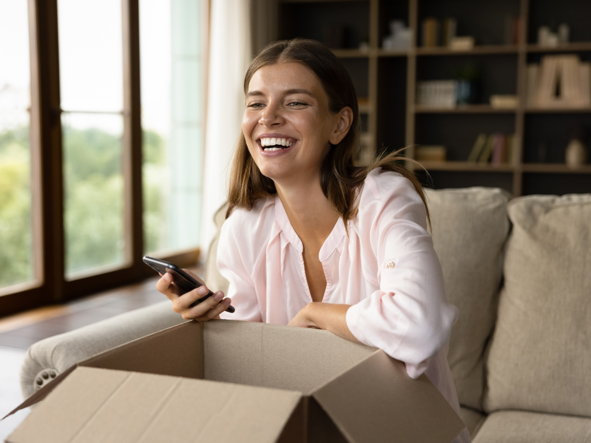 A woman smiles joyfully while holding her phone and unboxing a package, having ordered it online on a commerce website